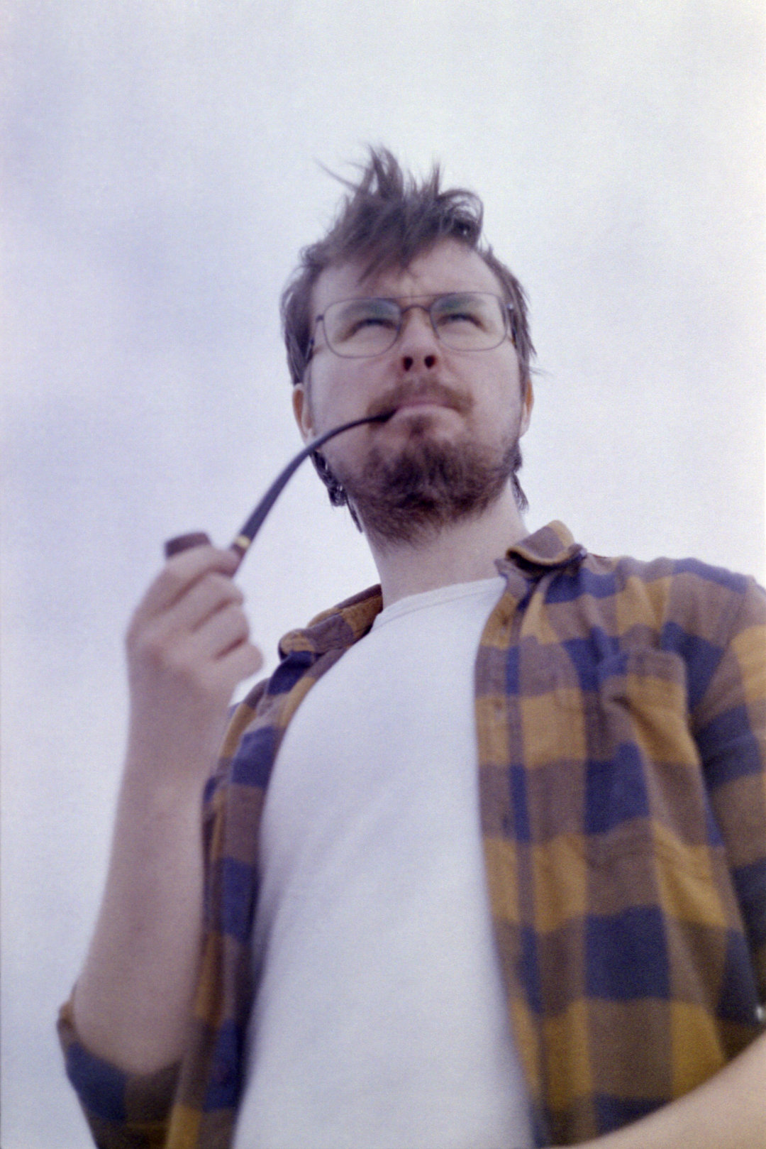 Photograph portraying a man in his early 30s taken midwaist slightly from below. He has a white t-shirt and a plaid shirt on top. He wears new retro glasses and smokes a churchwarden pipe. His hair is messy from the strong wind.