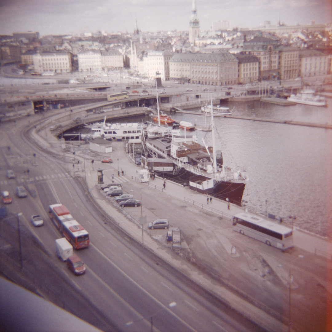 Photograph of the urban area Slussen in Stockholm with a couple of busses in traffic and one bigger boat docked to the harbour among some smaller ones further back. The horizon shows the cityscape of the citys old town.